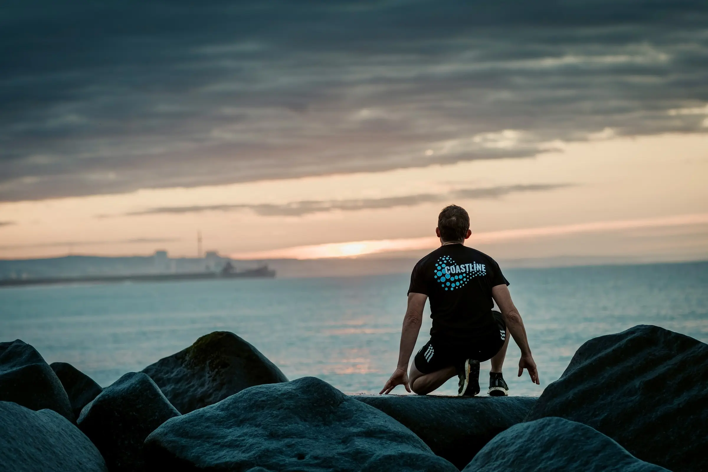 A man in a Coastline T-shirt crouches on the rocks, looking out across the sea towards the harbour.