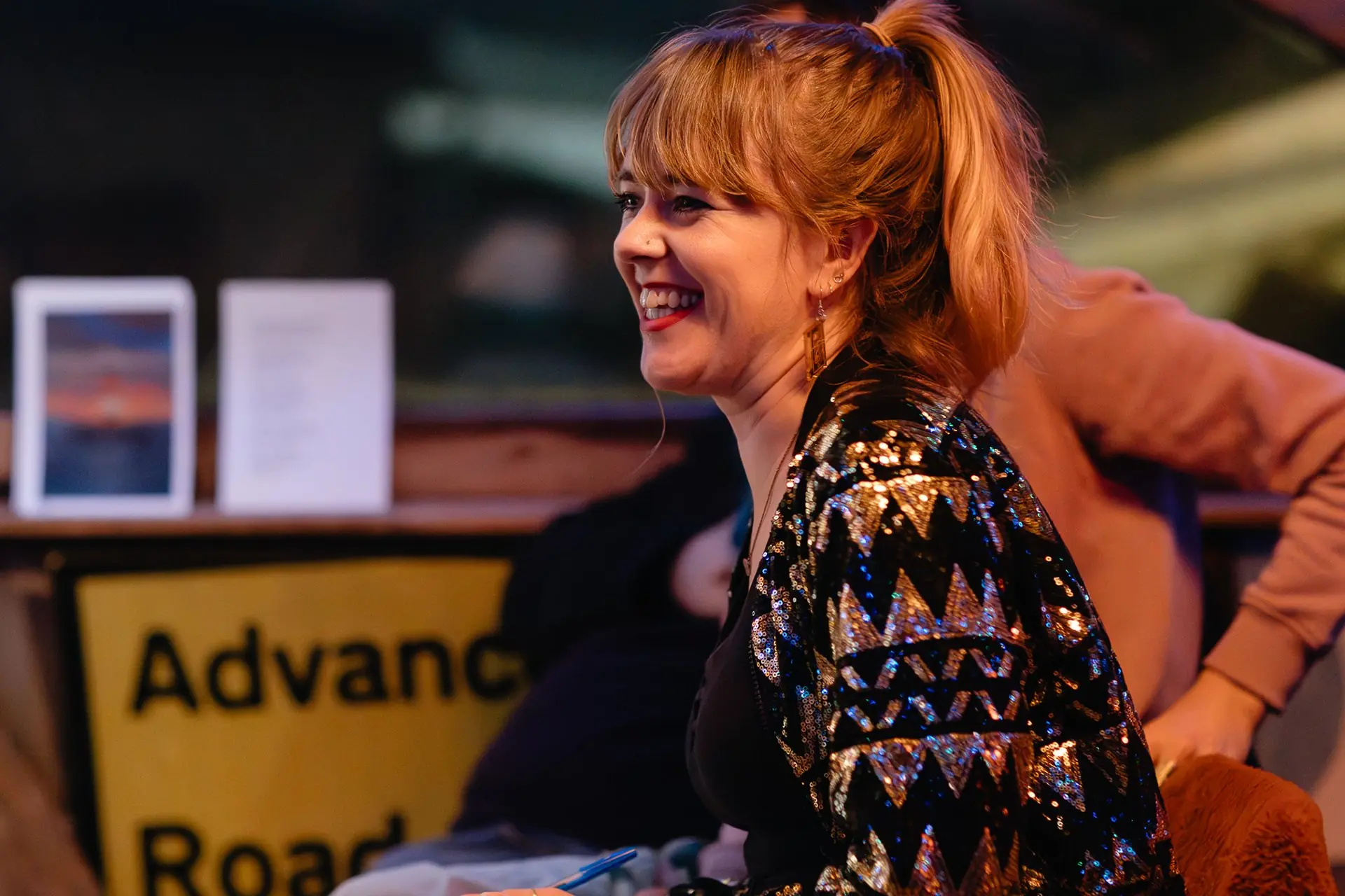 A woman in a sequinned jacket laughs beside a small table display.