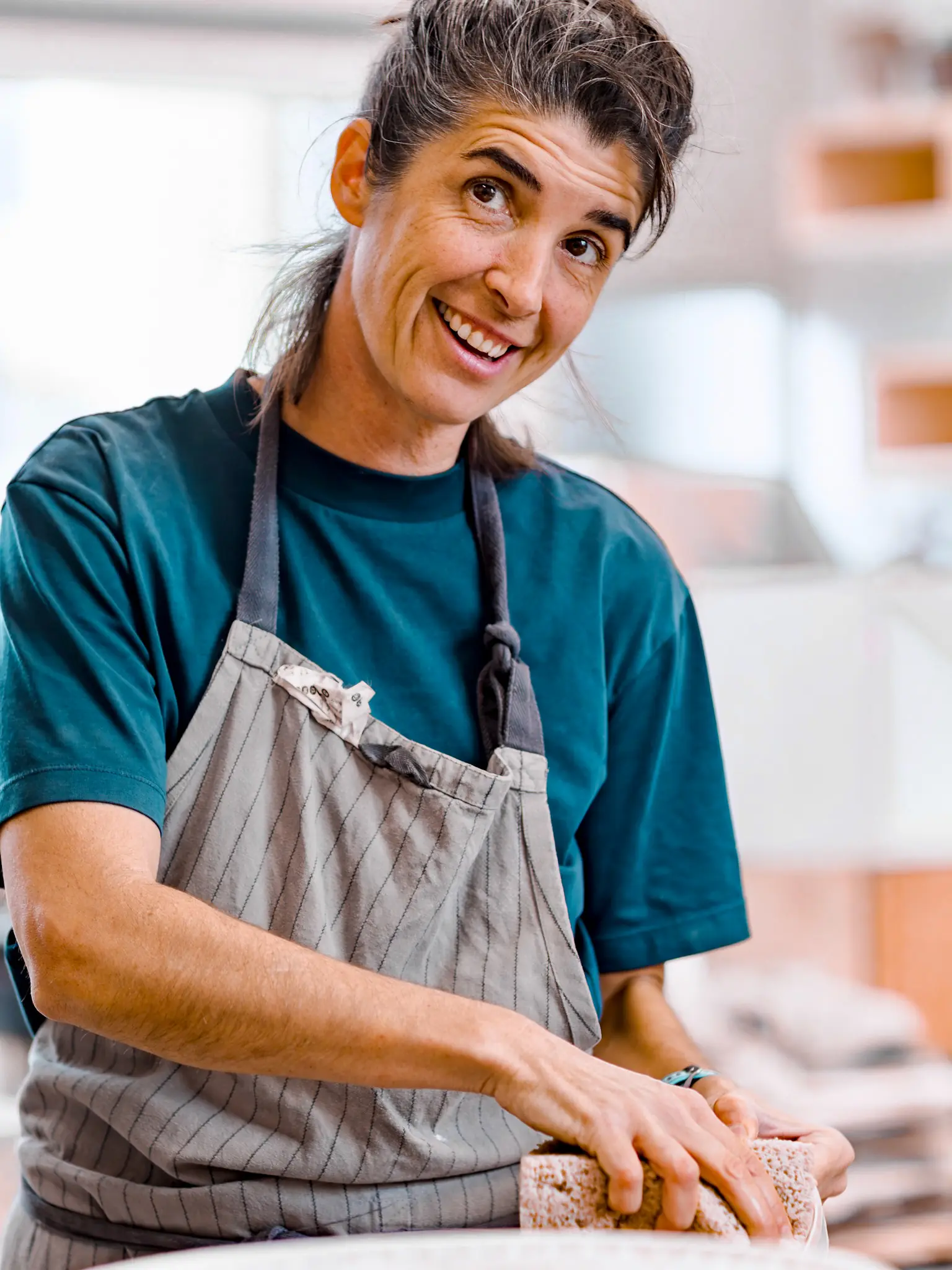 A potter in a teal T-shirt and grey apron smiles as she works clay at her bench.